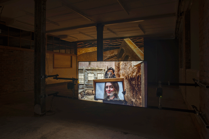 In a dark room a film is projected onto a screen. The projected image shows a light skinned person with dark hair and a facemask holding a tv screen with another light skinned, dark haired person with red lipstick smiling out from it