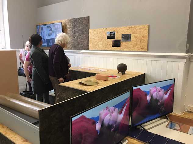 People starting at the exhibition, at images in corks. There are also two screens showing two people in red tops lying down heads agains each other.