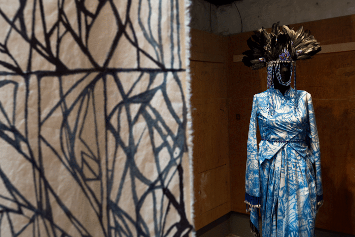 In the foreground a batic hanging on calico. In the background a manequin with an intricate dress made from fabric with an abstract indigo painted pattern. The manequin wears a headdress of large black feathers with strands of beads draped around the face.