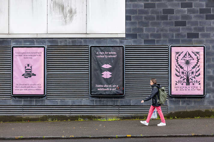 A row of posters are displayed outside on a dark building wall. A person walks past.