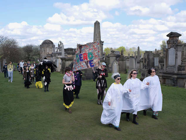 A procession in the Glasgow Necropolis lead by three women in white gowns. Behind them, people in balck and informal clothing. Some people are carrying a colorful banner that reads "not dead yet"