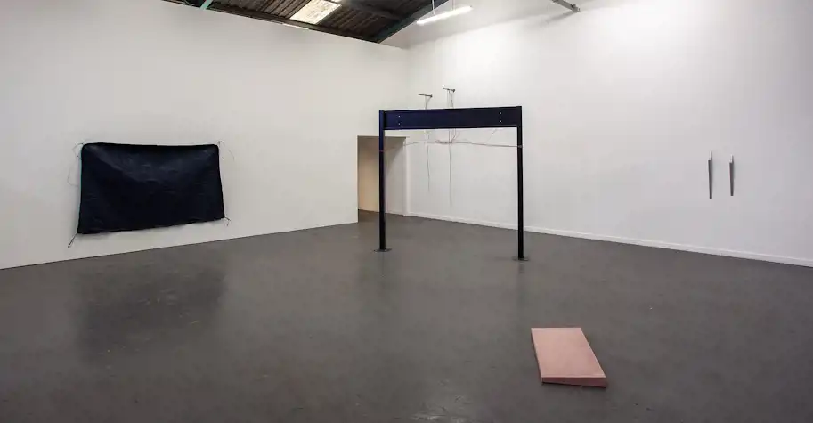An installation of abstract sculptures. There is a large black rectangle of fabric on the wall, a black metal structure standing in the space and a pink cast concrete rectangular shape with a small incline in its depth. On the right wall are two small cast aluminium abstract elongated shapes.