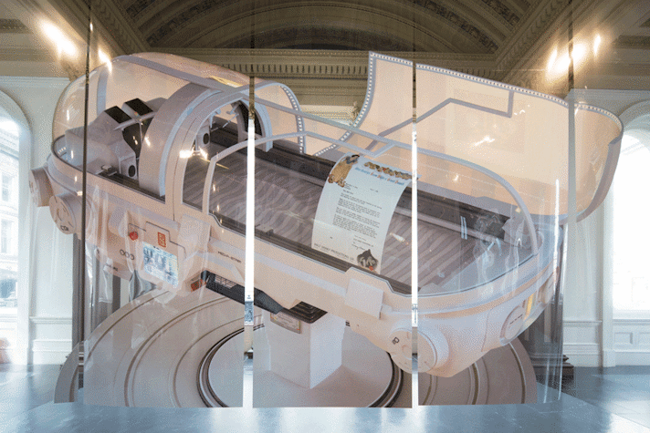 A futuristic looking mechanical, capsule is printed on a wall of polyester foil panels. It has a control screen on the side, a paper with a letter and an image of Snow White and the 7 Dwarves stuck on the glass of the capsule. The capsule is attached to a disk like base.