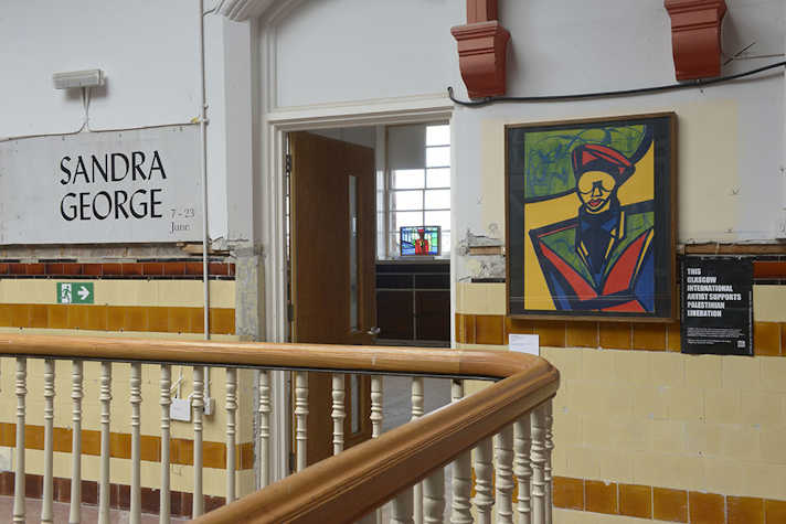 A view of the outside of the Sandra George Exhibition. There is a colourful artistic portrait of Sandra displayed on a wall and the words "Sandra George" on the wall next to a doorway. In the foreground is a bannister.