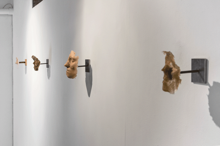 A white wall with several masks of faces made with different materials of warm/brown colours facing the public.