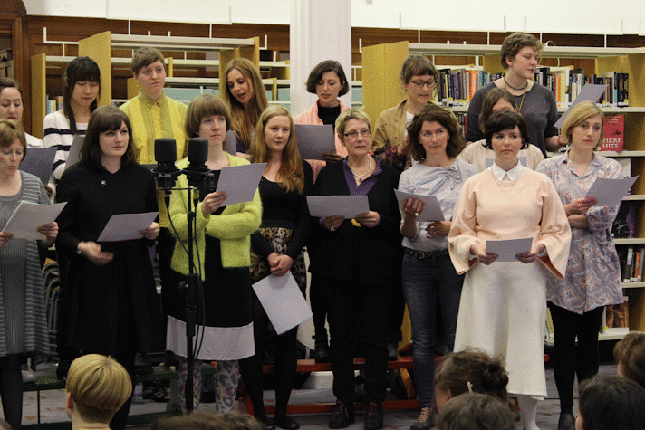 Two rows of women are standing in front of a seated crowd, They are holding pieces of paper and there are a couple microphones placed in front of them. Behind the women three are multiple bookshelves, carrying books.