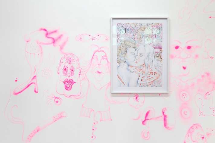 Framed drawing in colour pencil, depicting in fine detail 2 figures kissing, hung on a wall with abstracted pink figures and faces.