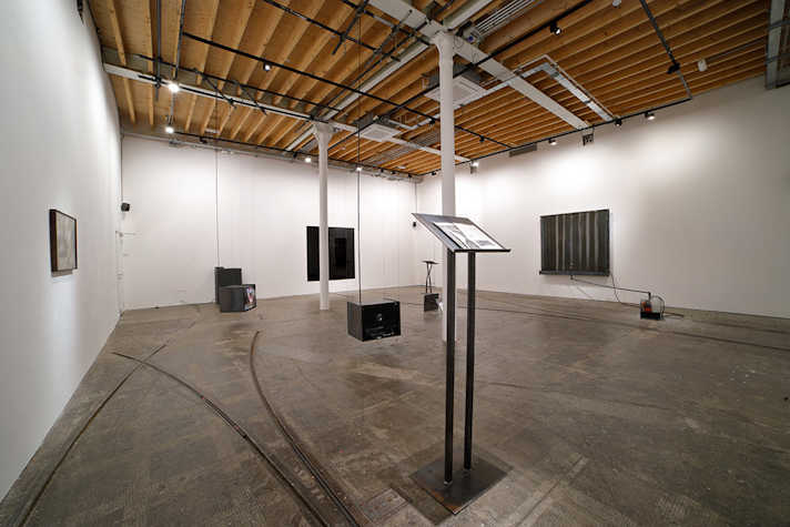 A white room which features artworks presented on walls and metal sculptures around the room, including a large metal lectern.