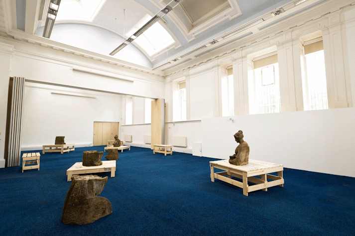In a large room with period feature ceiling and blue carpet, a series of wooden structures and sections of red sandstone sculptures are spaced out