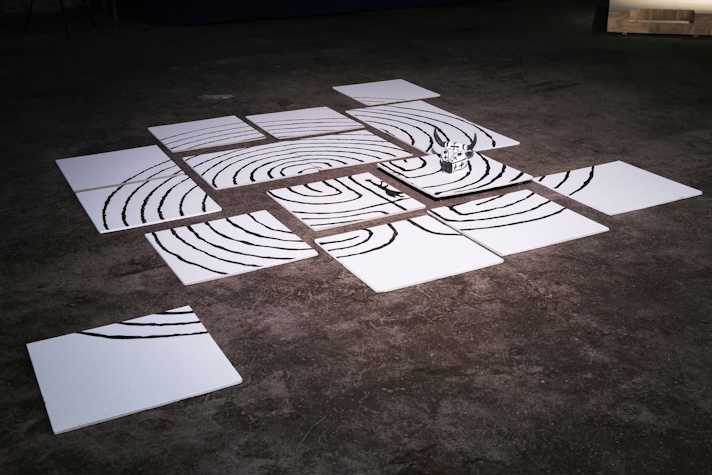 A series of white panels laid out on a floor. The panels have a series of black lines painted on them in concentric circles. A small paper model of a bull's head is also visible