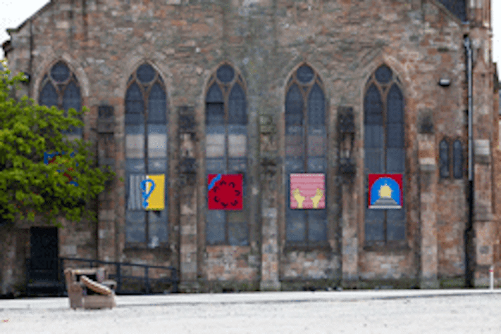 exterior of a church with four pictorial graphic flags hanging on each window. there is a singular arm chair in front of the church.