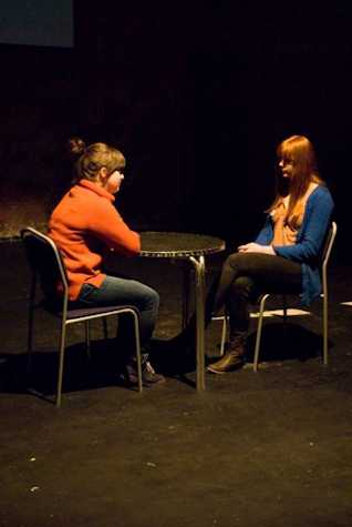 two people in an all black room sat on chairs facing each other with a table in between them