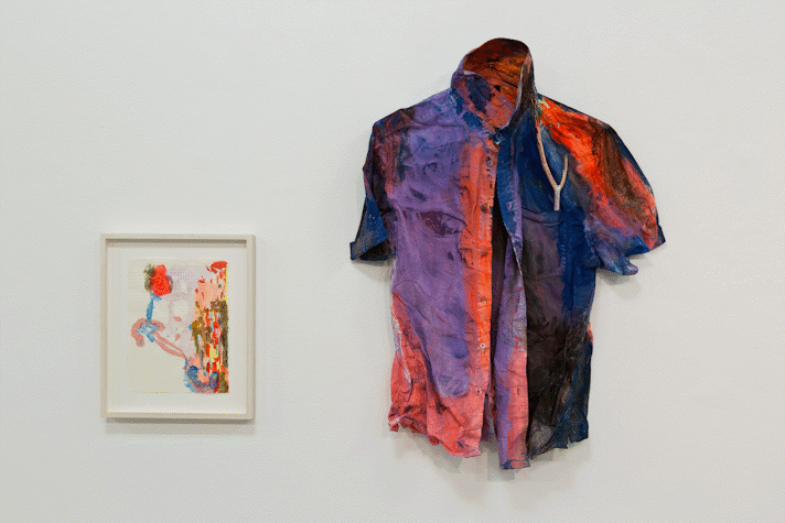 a pink shirt, crumpled and painted across in neon orange and purple, black and dark blue, hangs on a white wall next to an abstract painting on lined paper
