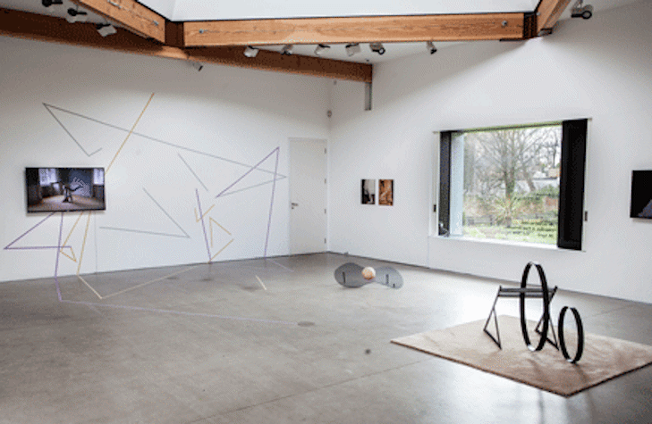 In a white studio, there are two different small sculptures made of metallic and plastic materials. There is a screen with a video of someone moving in the wall, which has colorful lines painted. There is a window on the right that looks out to a garden.
