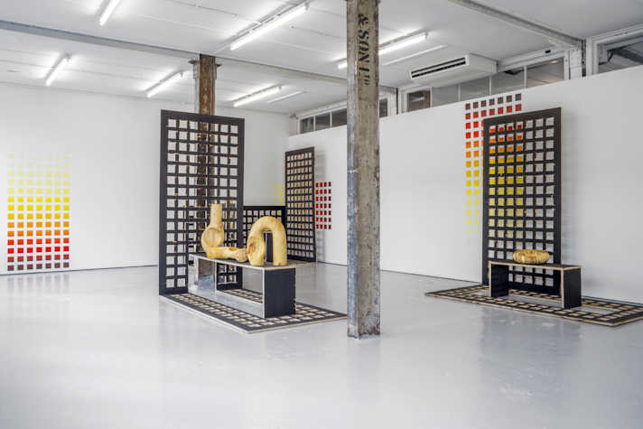 In a large room, there are multiple wooden sculptures and tall grid like structures placed around it. The walas have the negative space of the same grid painted on them. There are two iron pillars in the middle of the room and the artwork.