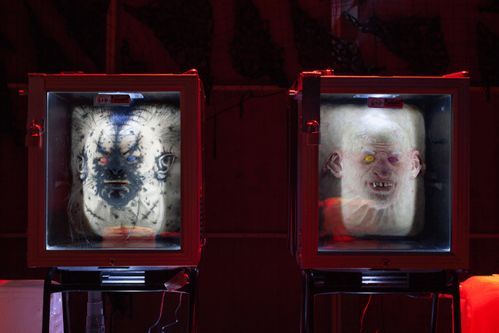 Two mangled half-rat half-human faces sit in mini-fridges under red light.