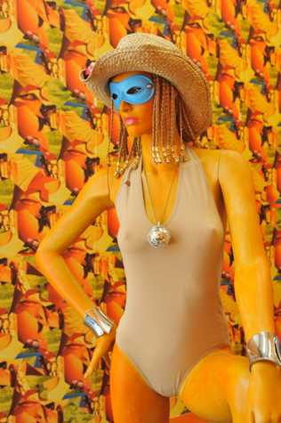 close up of a mannequin wearing a beige swimsuit, silver bangles and a disco ball necklace. the mannequin also dons a straw hat, blonde braids with beads at the end, and a blue eye mask. the background is a heavily patterned wallpapered wall.