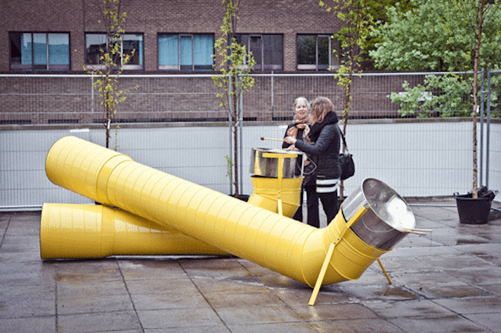 two yellow metal tubes with steel drums at the end of them. two women play with one of the drums.