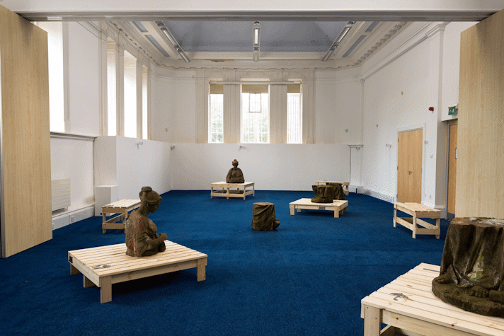 In a large room with period feature ceiling and blue carpet, a series of wooden structures and sections of red sandstone sculptures are spaced out