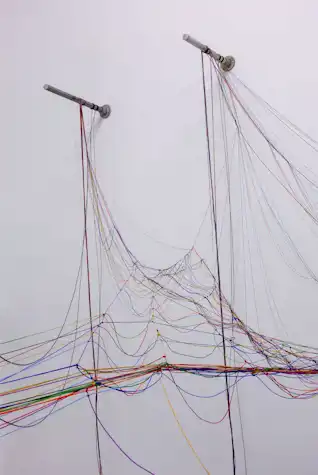 An abstract sculpture resembling a messy net of multicoloured cord hangs and branches down from two metal rods, mounted on a white wall.