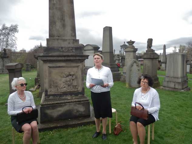 Performance in Glasgow Necropolis. Three women in white cardigans and black skirts and dark purses are sitting down and reading out loud from a paper with tomstones behind them.