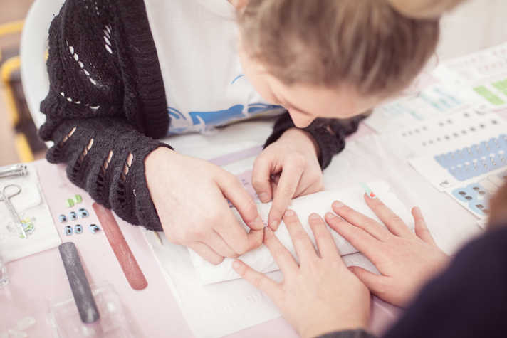 A woman is bent forward over a table, she is placing a nail art sticker on another person's hand. the table has a couple nail files, sticker packs and a pair of scissors placed in it.