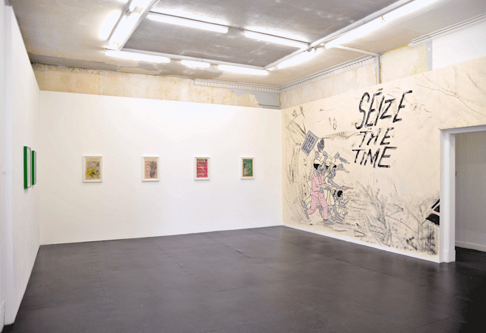 gallery with lithograph prints installed along the left and back walls. the right wall shows a mural with a group of protesters holding a picket sign and guns. mural text reads, 'seize the time'