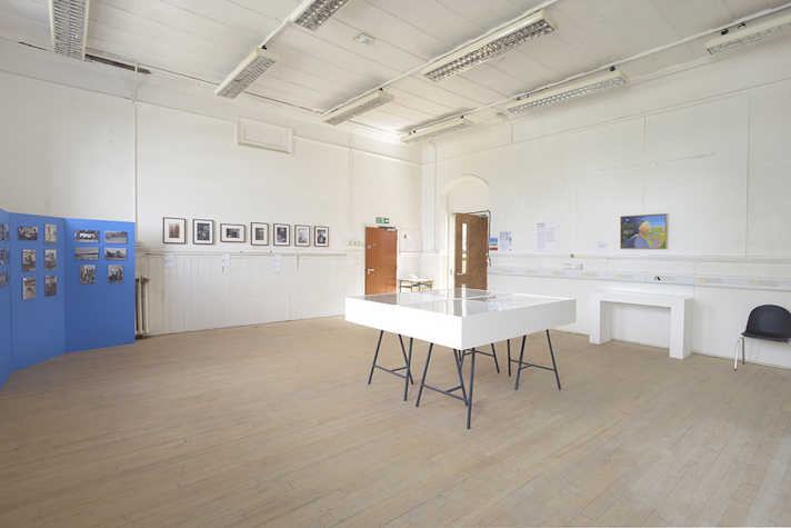 Photographs are displayed on the walls of a white room. In the centre there is a white table topped with glass.