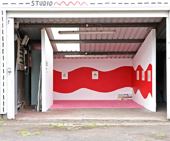 an open sided industrial space. Inside it 3 gallery walls frame a pink carpet. A red swiggle is painted across the walls and small framed drawings are hung on the wall