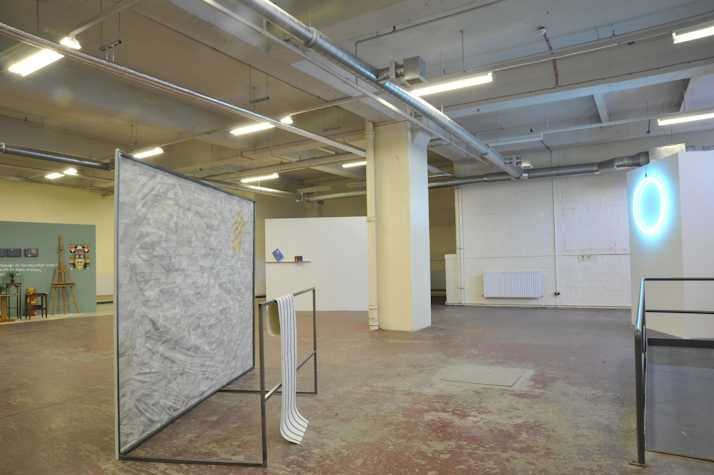installation of 'Petrosphere' group show. there is a cyan glowing neon circle in the back right. centre left shows a large metal freestanding frame with paper stretched across it. back right shows easels stools and plinths against a dark turquoise wall.