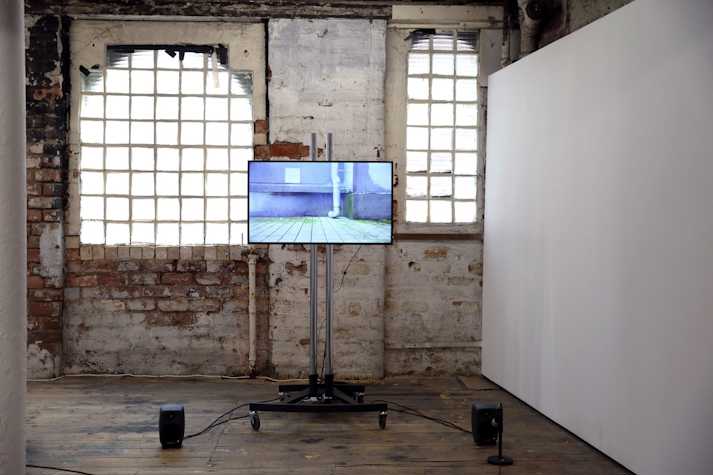 The image shows Yuko Mohri's installation in the pipe factory. A tv screen is on a stand and wired up to speakers on the floor. The screen shows a tiled floor and the bottom of a drainpipe.