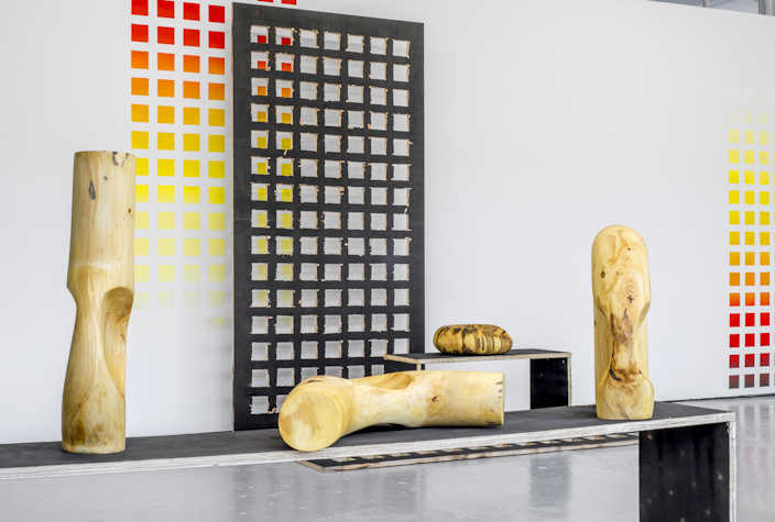 On a bench there are three wooden sculptures, behind which there is another round wooden sculpture on a smaller bench. There is a grid like structure against the walls and a grid like print on the wall.