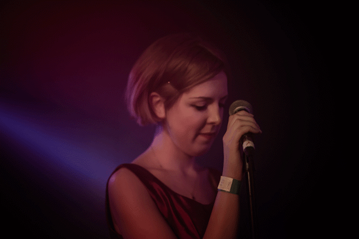 A woman is neatly dressed, with hair tucked behind her ear. She is holding a microphone with one hand, while looking in a downward direction.