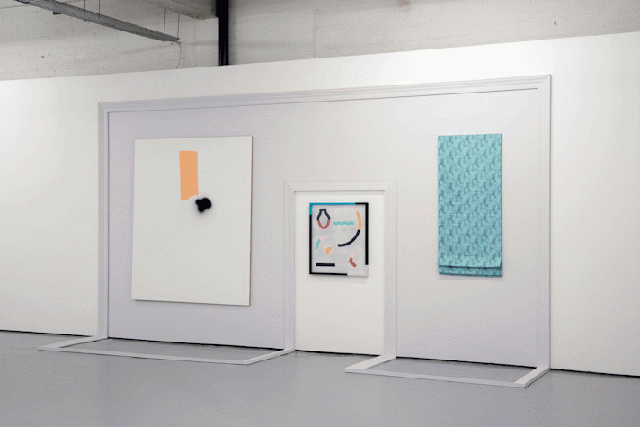 A large frame is made around a door, moving onto the floor. The frame has two artworks placed centrally on either sode of the door. One is a cloth and the other is like a painting. On the door, there is a multimedia artwork placed centrally in the top half of it.