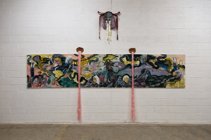 A wide rectangular painting on paper hangs on a white blockwork wall. There is an inky hint of a landscape, heads floating in space with links of sausages joining their mouths, a mountain has a snide face, hints towards hands, ghost figures, guts, clouds. Three, clay, mask-like faces hang above the painting, adorned with colourful hair. Two strands of pink hair hang down in front of the painting.