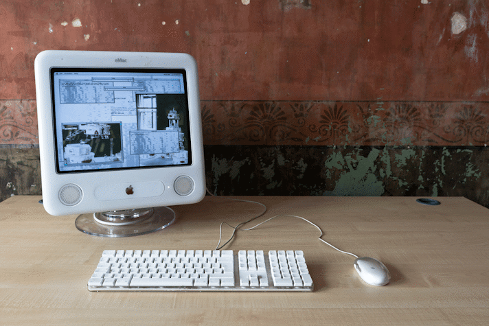 An early 2000s Mac computer sits on a beige laminate office desk with a keyboard and mouse. The screen has multiple different sized windows open, some with text some with glitchy black and white images.
