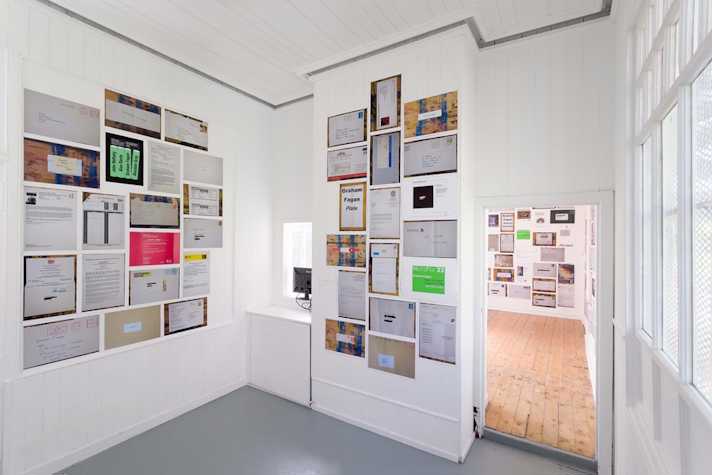 colourful photocopied sheets are pasted to the walls of a small gallery space. The sheets show letters, emails documents and other text-based media.