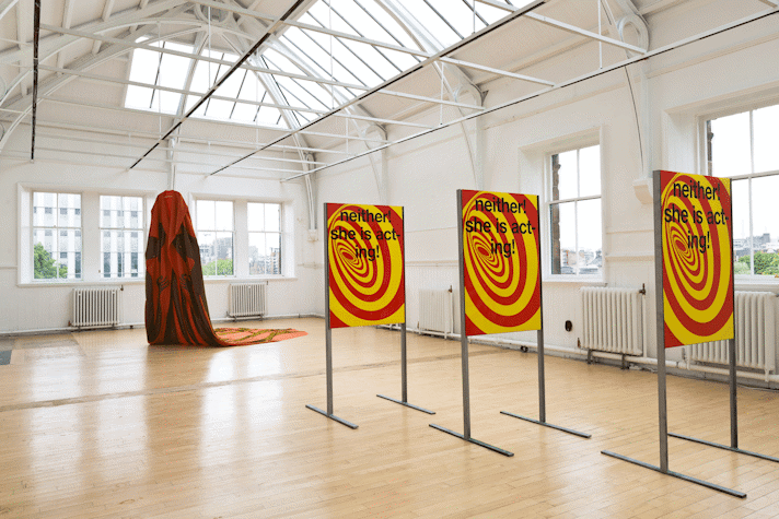Three identical metal stands hold three identical portrait oriented graphic posters with a red and yellow swirl and black text overlaid. The text reads 'neither! she is acting!'. In the backround a large cape is suspended from the roof and trails along the floor.