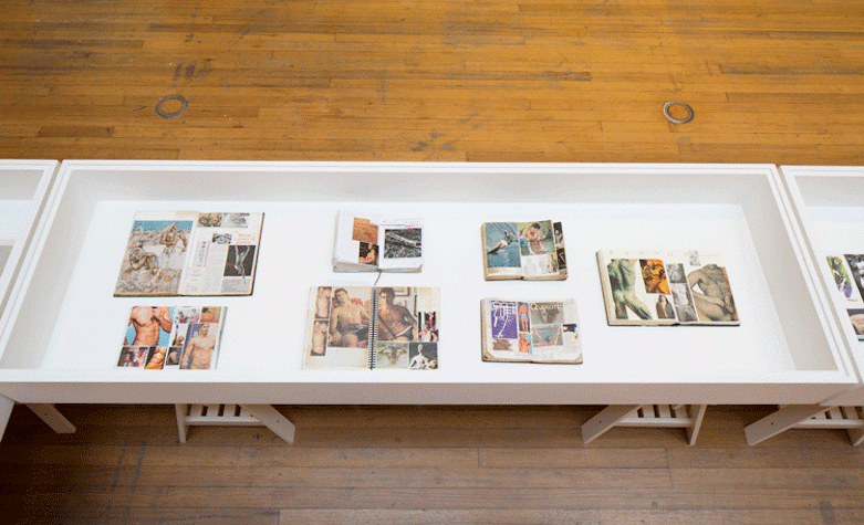 A low table is placed on a wooden floor, there are journal and pictures placed on this table. They are evenly space out. The pictures are from magazines and of men in various poses and settings. There are some men wearing underwear, some with a naked torso and one with the back image of a naked man. There is one man exercising and one man jumping. These journals are encased in glass, placed on top of the table.