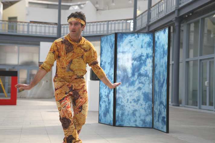 man wearing a suit and headband with images of pasta on it. he stands with knees slightly bent and arms outstretched. behind is a trifold screen with blue tie-dye/marble patterning.