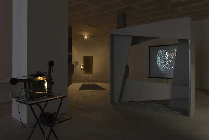 Darkened art gallery with a projected image of a man’s profile over a textured background, an old film projector on a stand, and various abstract installations in the background under soft lighting.