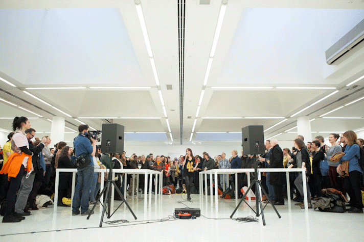 A woman is speaking into a microphone, standing in the centre of a large hall. There is a big crowd standing at a distance, all around her. In front of her, there are two long tables running parallel to each other. On the left there is a man taking a video of the event. Placed ahead of the tables and the man, are two speakers, held up by tripod stands. They are connected to a control box placed between them.