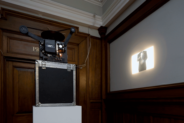 A projector is placed on top of a stack, consisting of a plinth and a box, projecting a small mid shot of a man in a suit onto a large projector screen. The shot of the man starts from his shoulders till his mid thigh. Thee walls of the room are wooden.