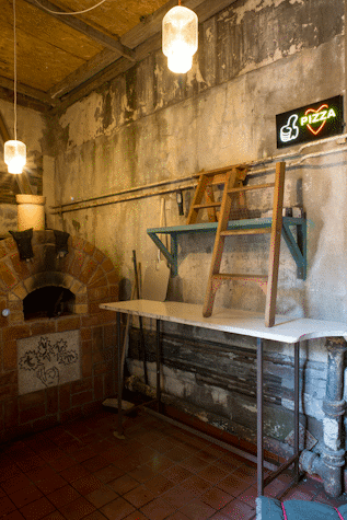 Cement room with two warm light lamps hanging from the ceiling. A black sign with a thumbs up with a Pizza enclosed in a heart. Some wood materials and tables are in the wall to the right. A stone oven is at the end of the room.