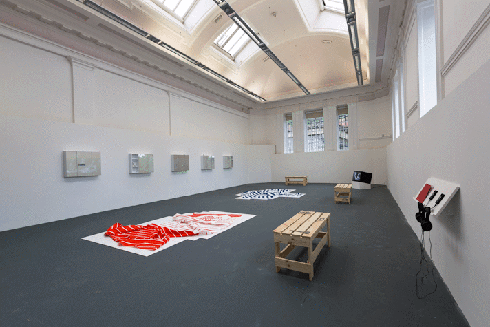 5 wall mounted cabinets, two floor sculptures, a TV, two benches, a shelf with headphones and publications are laid across a room