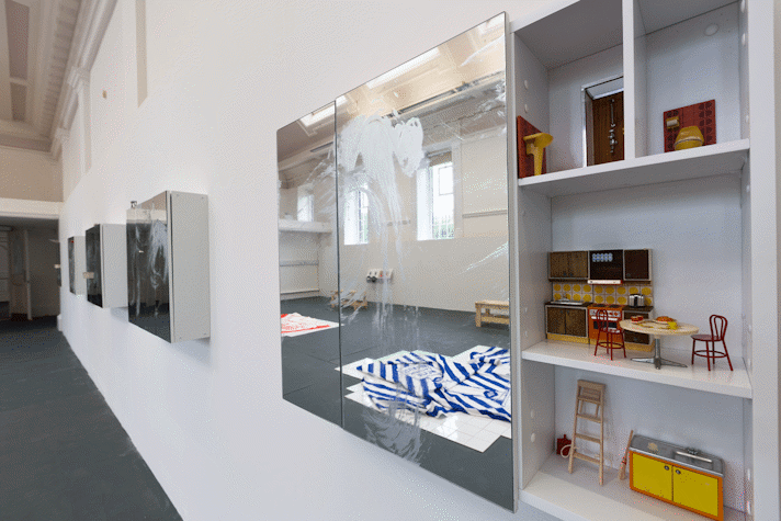 a mirrored cabinet mounted on a wall. One of the doors has been removed and reveals domestic scenes with dolls-house furniture inside.