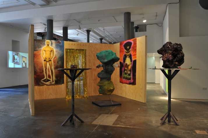 installation with plywood panels with a gold foil curtain opening. on the plywood panels there are two images pasted on and in front there are three sculptures