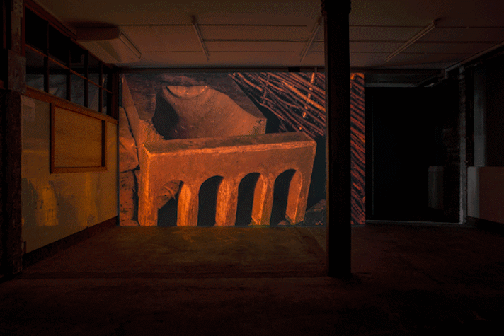 a dark industrial space with film projected onto a screen showing an abstract stone-like shape and wicker weaving