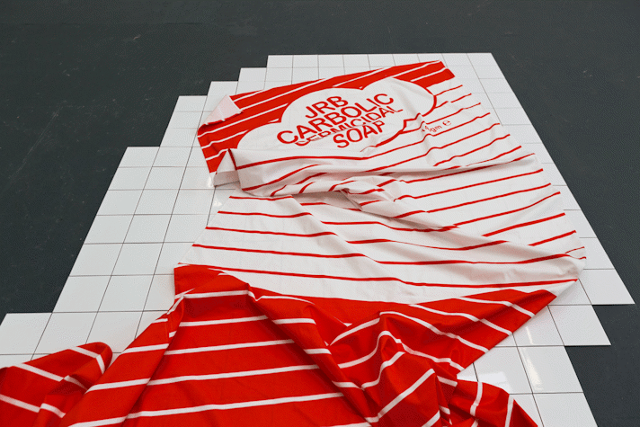 a red and white piece of striped fabric with printed lettering that reads 'JRB Carbolic Germicidal Soap' sits on a section of white tiled floor