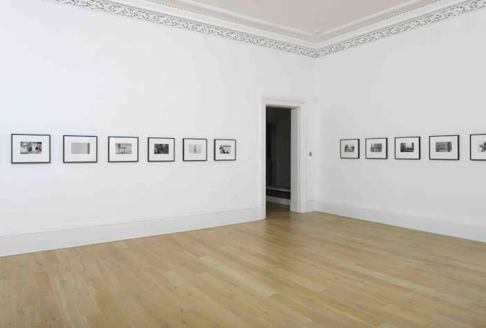 white gallery space with wooden floors. across the walls in a horizontal line are 11 images all in the same sized black frame.